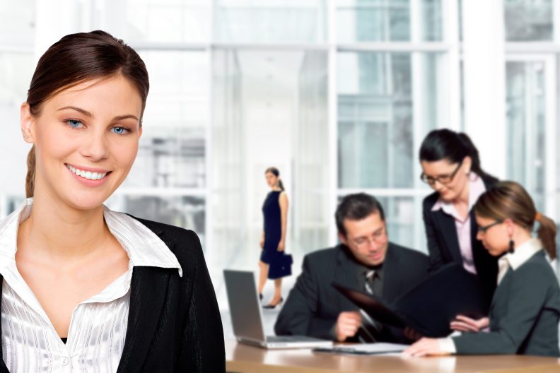 Eine lächelnde Geschäftsfrau im Vordergrund, während im Hintergrund ein Team von Kollegen in einem modernen Büro an einem Meeting arbeitet