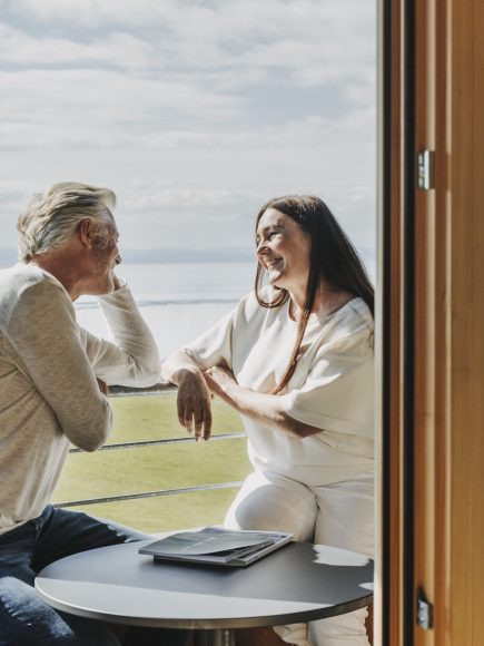 Ein Mann sitzt lächelnd auf einem Balkon und unterhält sich mit seiner Partnerin nach einer Andropause Behandlung.