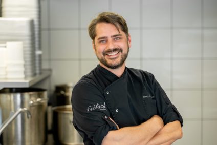 Ein lächelnder Koch in schwarzer Arbeitskleidung steht mit verschränkten Armen in einer modernen Küche