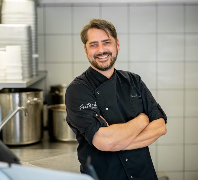 Ein lächelnder Koch in schwarzer Arbeitskleidung steht mit verschränkten Armen in einer modernen Küche