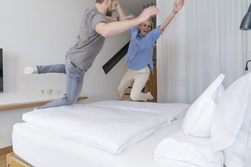 Ein lachendes Paar springt freudig auf einem großen Bett in einem modernen Hotelzimmer