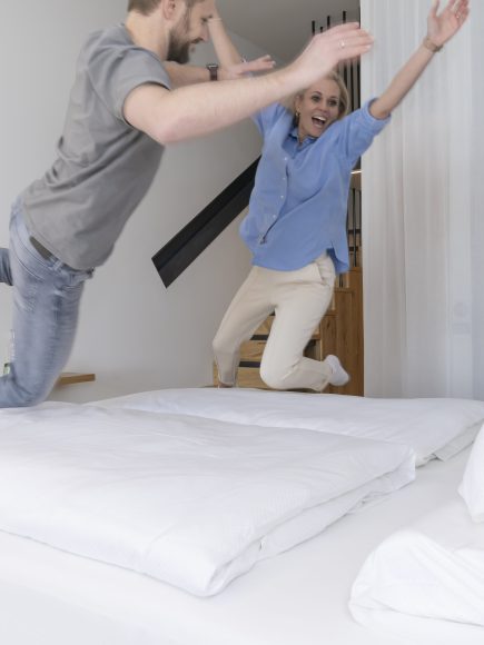 Ein lachendes Paar springt freudig auf einem großen Bett in einem modernen Hotelzimmer