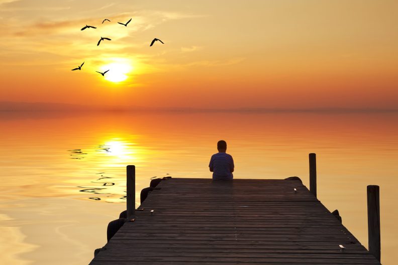 Eine Person sitzt auf einem Steg und blickt in einen farbenprächtigen Sonnenuntergang über einem ruhigen See, während Vögel am Himmel fliegen